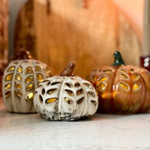 May include: Three ceramic pumpkin decorations in cream and orange. The pumpkins have cut-out designs that allow light to shine through, creating a warm glow. The pumpkins are arranged on a white surface, ideal for autumn decor.
