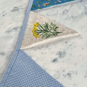 May include: A handmade bunting banner with three triangular flags. The top flag is blue with a floral pattern, the middle flag is beige with yellow embroidered flowers, and the bottom flag is blue and white gingham. The flags are connected with a blue and white gingham trim.