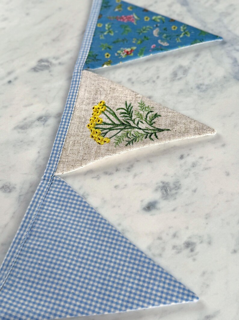 May include: A handmade bunting banner with three triangular flags. The top flag is blue with a floral pattern, the middle flag is beige with yellow embroidered flowers, and the bottom flag is blue and white gingham. The flags are connected with a blue and white gingham trim.