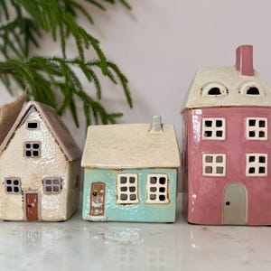 Peut inclure: Trois figurines de maison en c&eacute;ramique cr&egrave;me, turquoise et rose. Chaque maison a un design diff&eacute;rent, avec des fen&ecirc;tres et des portes. Les maisons sont d&eacute;coratives et conviennent &agrave; la d&eacute;coration int&eacute;rieure. Les maisons sont de tailles variables.