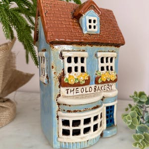 Può includere: Una miniatura in ceramica di una panetteria, dipinta di azzurro chiaro con tetto marrone. Le finestre sono bianche e l'insegna recita "THE OLD BAKERY". Fiori gialli nei vasi. Un oggetto decorativo.