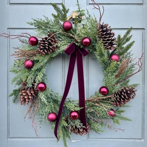 May include: A Christmas wreath made of evergreen branches, pine cones, and burgundy baubles. A burgundy velvet ribbon is tied around the wreath.