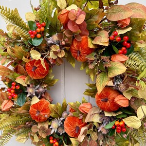 May include: A decorative wreath featuring orange velvet pumpkins, clusters of bright red berries, and an assortment of green, yellow, and brown autumn leaves. The arrangement includes dried thistle accents and various foliage, creating a dense, circular display of seasonal textures and colors.