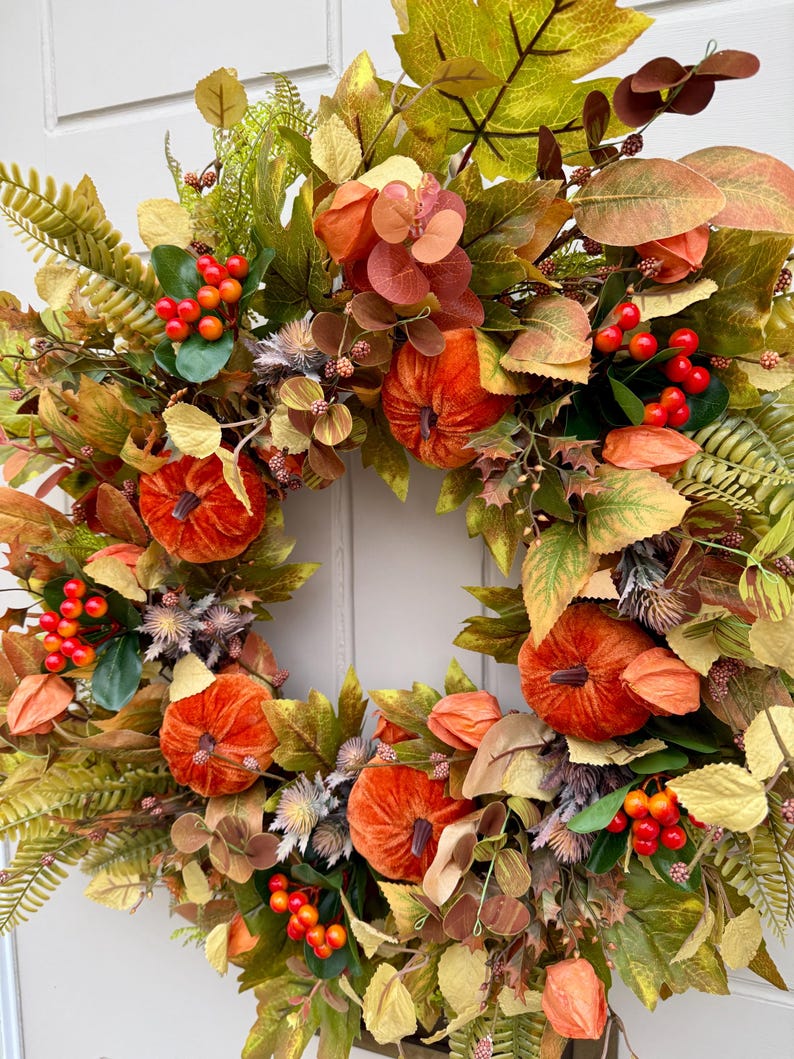 May include: A decorative wreath featuring orange velvet pumpkins, clusters of bright red berries, and an assortment of green, yellow, and brown autumn leaves. The arrangement includes dried thistle accents and various foliage, creating a dense, circular display of seasonal textures and colors.