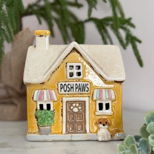 May include: A ceramic house ornament with a yellow facade and a cream roof. The sign reads "POSH PAWS" above a brown door with a paw print. A small dog figurine sits to the right. The house has two windows and two awnings.