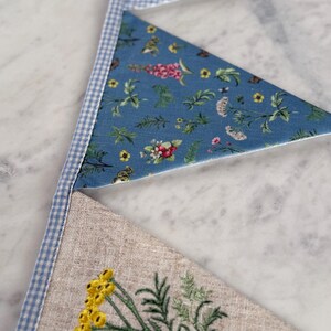 May include: Close-up of a handmade bunting with three triangular flags. The top flag is blue and white gingham, the middle flag is blue with a floral pattern, and the bottom flag is beige linen with yellow embroidered flowers and green leaves.
