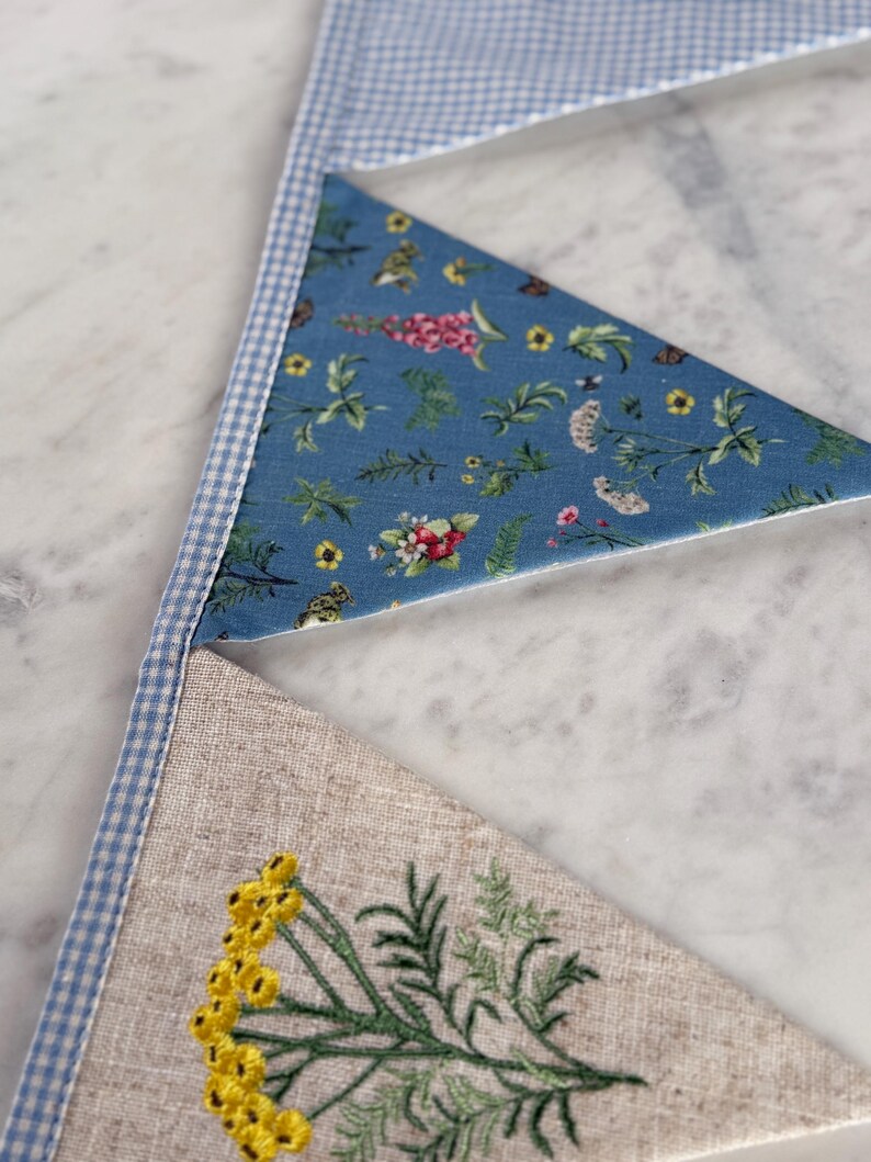 May include: Close-up of a handmade bunting with three triangular flags. The top flag is blue and white gingham, the middle flag is blue with a floral pattern, and the bottom flag is beige linen with yellow embroidered flowers and green leaves.