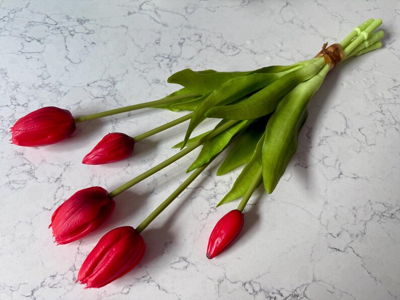 May include: A bouquet of red tulips with green stems and leaves, tied together with a brown twine. The vibrant red petals contrast with the green foliage, creating a fresh and natural aesthetic. The flowers are arranged on a white marble surface.