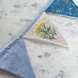 May include: A handmade bunting banner with triangular flags. The flags feature various patterns, including blue gingham, floral prints, and a linen flag embroidered with yellow flowers and green leaves. The banner is displayed on a marble surface.