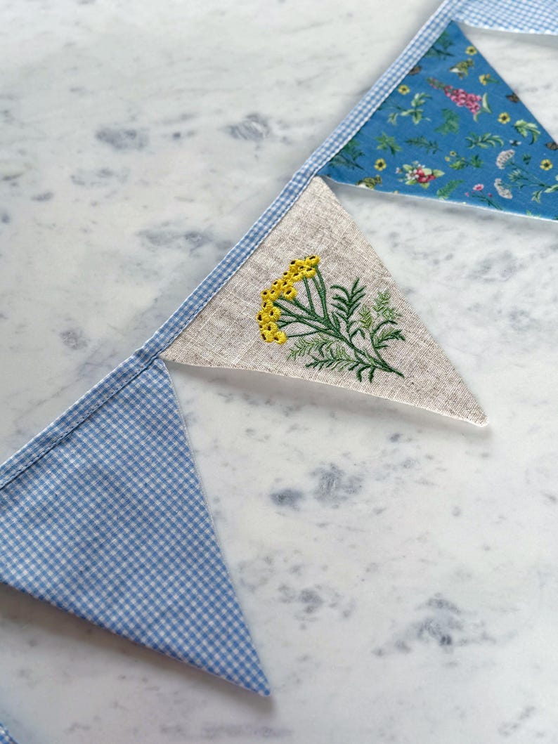 May include: A handmade bunting banner with triangular flags. The flags feature various patterns, including blue gingham, floral prints, and a linen flag embroidered with yellow flowers and green leaves. The banner is displayed on a marble surface.