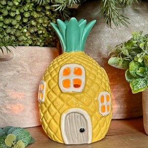 May include: A yellow pineapple-shaped ceramic house with a green leafy top. The exterior features a textured diamond pattern, a grey arched door with a black knob, and three white-framed windows glowing with a warm orange light from within.