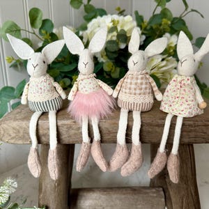 Könnte beinhalten: Vier weiße Filz-Hasenfiguren mit langen Beinen, die auf einem Holzhocker sitzen. Jeder Hase trägt ein anderes gemustertes Kleid: grün-weiß geblümt, rosa-weiß geblümt, braunes Gingham und weiß-rosa geblümt.