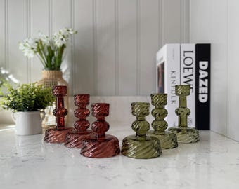 May include: Set of five glass candle holders, three in a deep burgundy color and two in a muted olive green. The candle holders have a twisted design and are arranged on a white surface.
