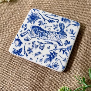 May include: A white ceramic coaster with a blue and white design of a rabbit running through a field of flowers and plants.