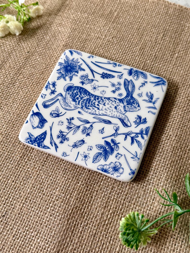 May include: A white ceramic coaster with a blue and white design of a rabbit running through a field of flowers and plants.