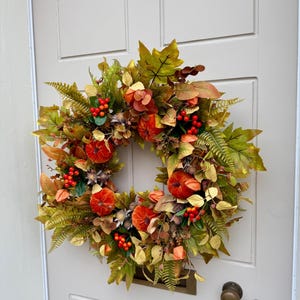 May include: A circular autumn wreath featuring artificial orange pumpkins, green and yellow maple leaves, fern fronds, and clusters of small red berries. The arrangement is dense with varied foliage, incorporating dried-look textures and warm seasonal colors.