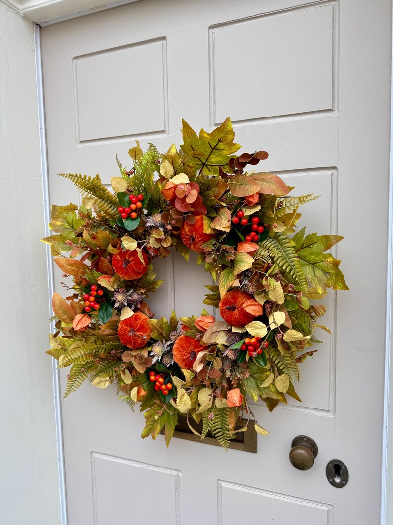 May include: A circular autumn wreath featuring artificial orange pumpkins, green and yellow maple leaves, fern fronds, and clusters of small red berries. The arrangement is dense with varied foliage, incorporating dried-look textures and warm seasonal colors.