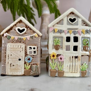 May include: Two ceramic house ornaments. One is grey with a heart-shaped window and a door. The other is white with a heart-shaped window and a door. Both have flower and plant details, and decorative bunting.