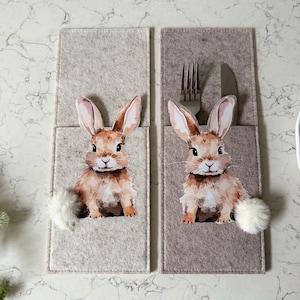 May include: Two beige felt place card holders with a bunny rabbit design and a pocket for cutlery. Each holder has a white fluffy pom-pom.