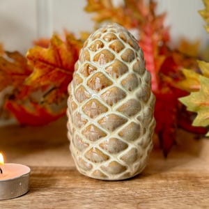 Puede incluir: Un objeto decorativo de cerámica con forma de piña, con una superficie texturizada y un acabado brillante. La piña es de color marrón claro con una base color crema. Una vela de té encendida está frente a la piña, con hojas de otoño en el fondo.
