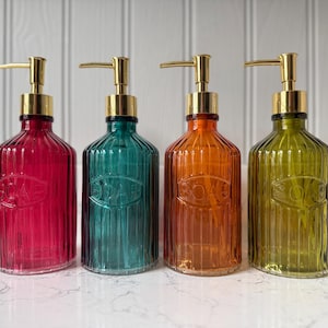 May include: Four coloured glass soap dispensers with gold-coloured pumps. The dispensers are in shades of red, teal, orange, and green. Each bottle has a ribbed design and the word "SOAP" embossed on the front.