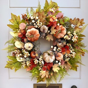 May include: A circular autumn wreath featuring an assortment of velvet-textured pumpkins in shades of orange, cream, and tan, interspersed with green and brown maple leaves, fern fronds, and clusters of small white and cream-colored berries.