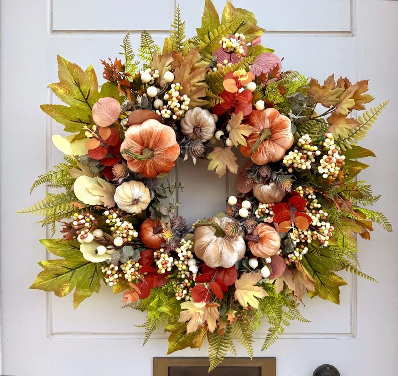 May include: A circular autumn wreath featuring an assortment of velvet-textured pumpkins in shades of orange, cream, and tan, interspersed with green and brown maple leaves, fern fronds, and clusters of small white and cream-colored berries.