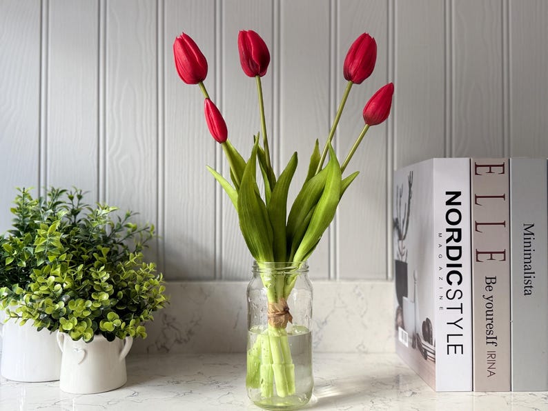 May include: A clear glass jar holds a bouquet of red tulips with green stems and leaves. The jar is tied with twine and sits on a white countertop. Decorative books and a small potted plant are in the background.