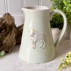 May include: A light green ceramic pitcher with a white lamb design. The pitcher has a wide mouth and a handle.