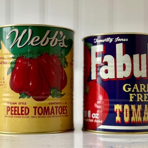 May include: Two vintage style metal cans of peeled tomatoes. The can on the left is yellow with green and red accents. The can on the right is blue with white, red, and gold accents. The can on the left reads 'Webb's Italian Style Peeled Tomatoes' and the can on the right reads 'Fabulos Garden Fresh Tomatoes'.
