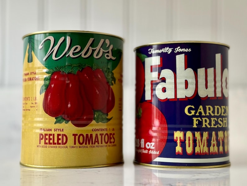 May include: Two vintage style metal cans of peeled tomatoes. The can on the left is yellow with green and red accents. The can on the right is blue with white, red, and gold accents. The can on the left reads 'Webb's Italian Style Peeled Tomatoes' and the can on the right reads 'Fabulos Garden Fresh Tomatoes'.