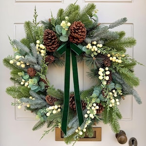 May include: A circular wreath featuring green pine branches, frosted evergreen needles, and brown pinecones. The arrangement is accented with clusters of small cream-coloured berries and eucalyptus leaves, finished with a long dark green velvet ribbon.