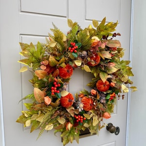May include: A circular autumn wreath featuring orange velvet pumpkins, clusters of bright red berries, and a variety of green, yellow, and brown faux leaves. The arrangement includes fern-like foliage and small dried seed pods, creating a dense, textured display of seasonal colors.