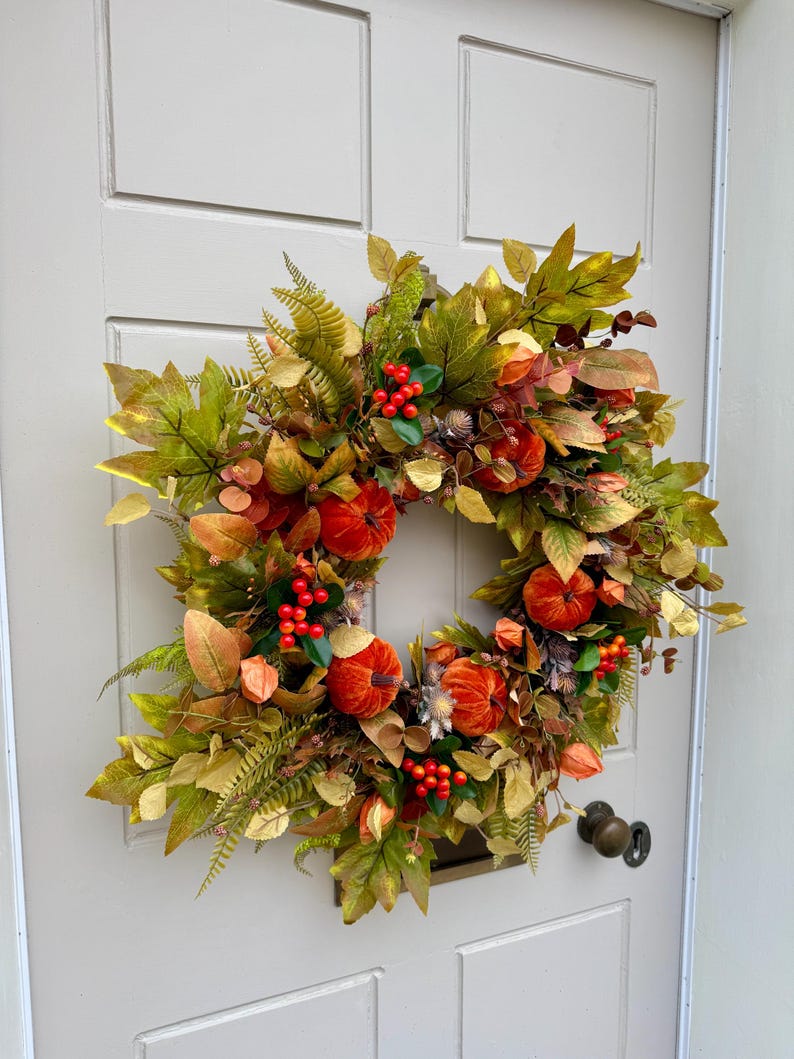 May include: A circular autumn wreath featuring orange velvet pumpkins, clusters of bright red berries, and a variety of green, yellow, and brown faux leaves. The arrangement includes fern-like foliage and small dried seed pods, creating a dense, textured display of seasonal colors.