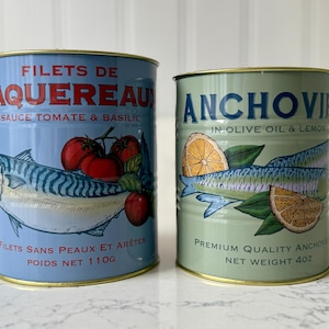 May include: Two canned fish products. One is light blue with a mackerel illustration, tomatoes, and text in French. The other is green with an anchovy illustration, lemon slices, and English text. Net weight of the blue can is 110g, and the green can is 4oz.