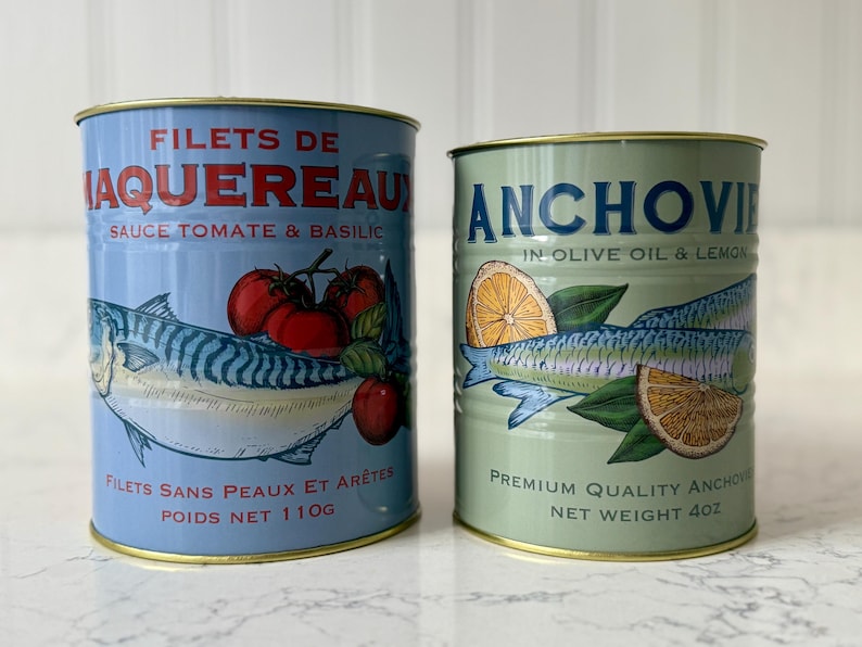 May include: Two canned fish products. One is light blue with a mackerel illustration, tomatoes, and text in French. The other is green with an anchovy illustration, lemon slices, and English text. Net weight of the blue can is 110g, and the green can is 4oz.