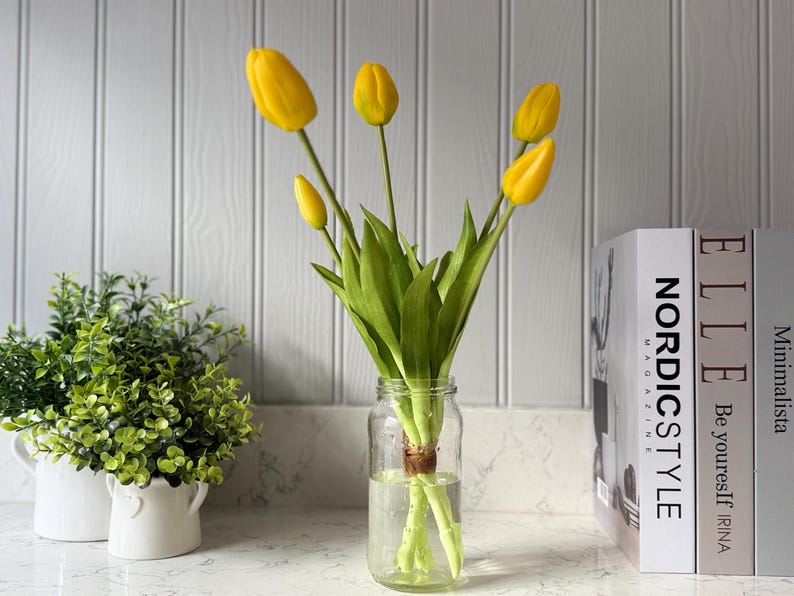 May include: A clear glass jar holds a bouquet of yellow tulips with green stems and leaves. The tulips are arranged in a vase with water. Two small white pots with green plants are on the left. Books with the titles "NORDIC STYLE" and "ELLE" are on the right.