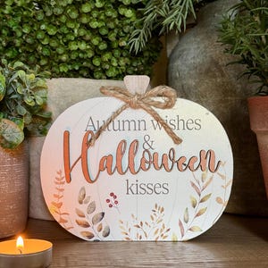 May include: A flat, pumpkin-shaped wooden sign featuring the text Autumn wishes & Halloween kisses in orange and grey lettering. The design includes a printed botanical pattern of leaves and berries, topped with a decorative jute twine bow.
