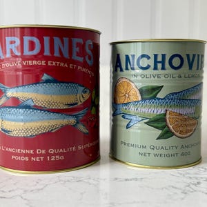 May include: Two cans of preserved seafood. One is red with the word "SARDINES" and fish illustrations. The other is green with "ANCHOVIES" and lemon slices. The red can's net weight is 125g, and the green can's net weight is 4oz.