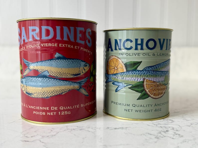 May include: Two cans of preserved seafood. One is red with the word "SARDINES" and fish illustrations. The other is green with "ANCHOVIES" and lemon slices. The red can's net weight is 125g, and the green can's net weight is 4oz.