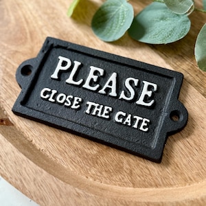 Op de afbeelding: Een rechthoekig zwart metalen bord met de witte, verhoogde tekst "PLEASE CLOSE THE GATE". Het bord heeft afgeronde hoeken en gaten voor bevestiging. Het ligt op een houten oppervlak.