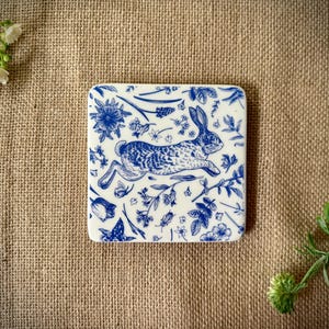 May include: A white ceramic coaster featuring a blue and white design of a rabbit running through a field of flowers.