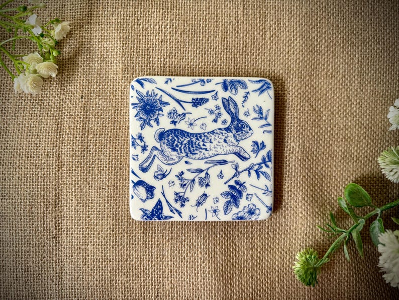May include: A white ceramic coaster with a blue and white design of a rabbit running through a field of flowers.