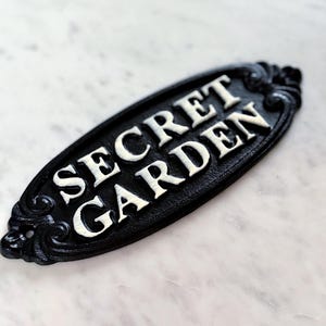 以下が含まれることがあります： 白い文字で「SECRET GARDEN」と書かれた黒い楕円形のサイン。サインの端には装飾的なスクロールワークがあります。背景は明るい大理石の表面です。