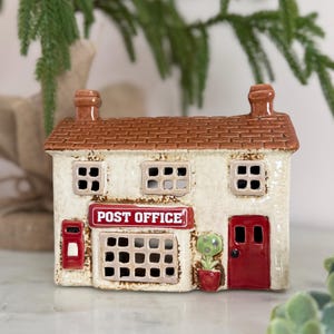 以下が含まれることがあります： 茶色の瓦屋根、クリーム色の壁、赤いドアを備えたセラミック製のミニチュア郵便局。看板には赤字で「POST OFFICE」と書かれています。小さな赤い郵便ポストと鉢植えの植物も見えます。