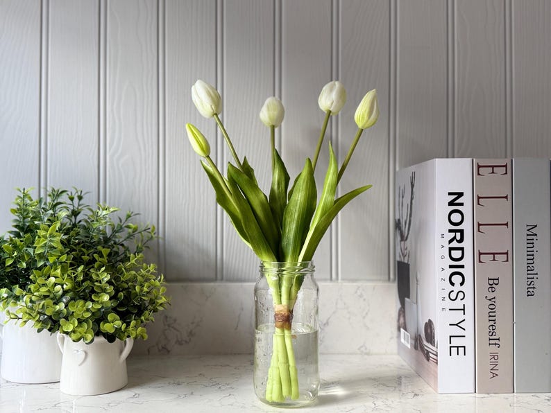 May include: A clear glass jar holds a bundle of white tulips with green stems and leaves. The jar is tied with twine and sits on a white countertop. Green leafy plants in white pots are to the left, and books are to the right.