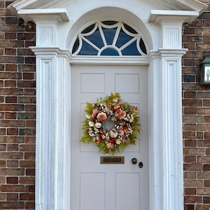 May include: A round autumn wreath featuring artificial pumpkins, berries, and assorted green and orange foliage. The wreath is mounted on a light gray paneled door with a brass mail slot and a circular door handle, framed by white architectural trim.