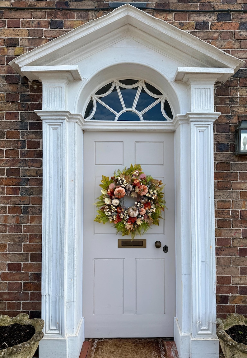 May include: A round autumn wreath featuring artificial pumpkins, berries, and assorted green and orange foliage. The wreath is mounted on a light gray paneled door with a brass mail slot and a circular door handle, framed by white architectural trim.