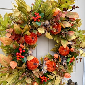 May include: A decorative autumn wreath featuring velvet-textured orange pumpkins, clusters of bright red berries, and a variety of green, yellow, and brown faux foliage including maple leaves and ferns, arranged in a circular shape on a white door.