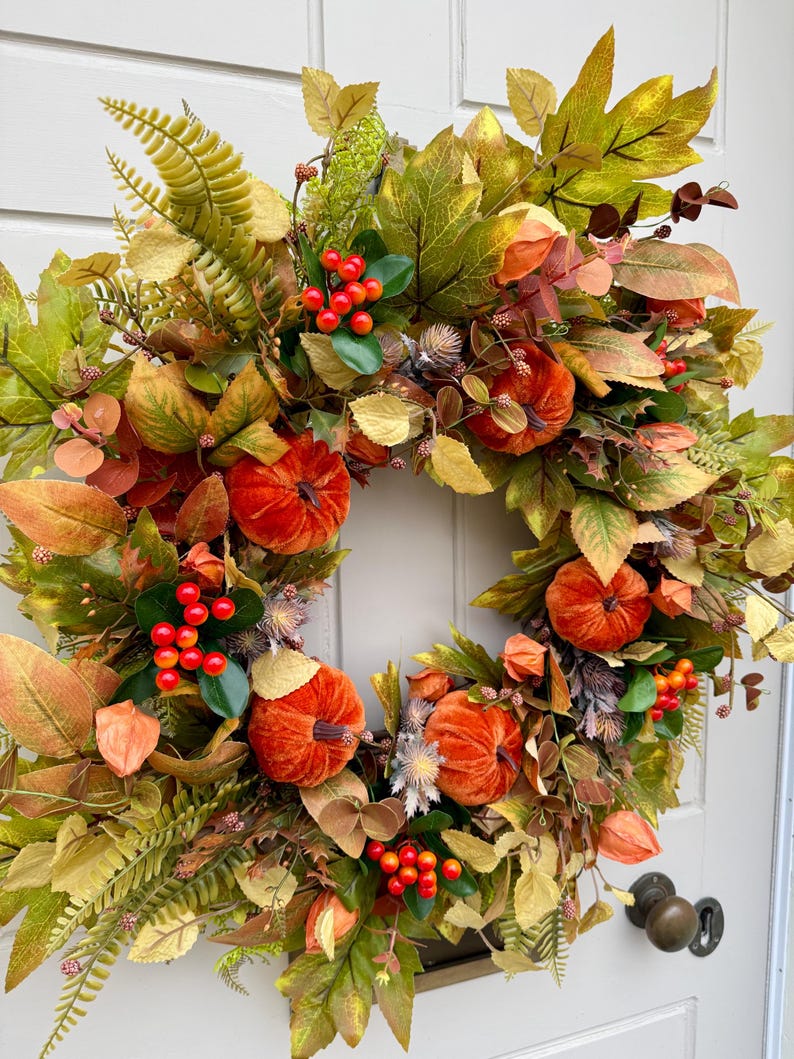 May include: A decorative autumn wreath featuring velvet-textured orange pumpkins, clusters of bright red berries, and a variety of green, yellow, and brown faux foliage including maple leaves and ferns, arranged in a circular shape on a white door.